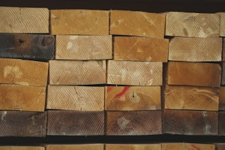 A neatly stacked pile of wooden planks, each showing distinct grain patterns and some with natural knots. The wood has a range of lighter to darker brown hues, with visible growth rings on the end surfaces. A few planks have dark spots and there are red marks on some surfaces.