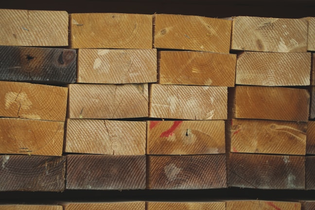 A neatly stacked pile of wooden planks, each showing distinct grain patterns and some with natural knots. The wood has a range of lighter to darker brown hues, with visible growth rings on the end surfaces. A few planks have dark spots and there are red marks on some surfaces.