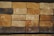 A neatly stacked pile of wooden planks, each showing distinct grain patterns and some with natural knots. The wood has a range of lighter to darker brown hues, with visible growth rings on the end surfaces. A few planks have dark spots and there are red marks on some surfaces.