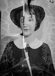 A black and white portrait of a person wearing a hat, with visible damage and cracks across the image. The photograph appears to have been overlaid with abstract textures and lines, giving it an aged and distorted appearance.