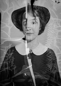 A black and white portrait of a person wearing a hat, with visible damage and cracks across the image. The photograph appears to have been overlaid with abstract textures and lines, giving it an aged and distorted appearance.