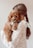 A young woman with long brown hair styled in a braid is holding a small, fluffy brown puppy. The puppy has curly fur and is looking towards the viewer with a gentle expression. The woman is wearing a knitted sweater with small colorful specks.