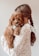 A young woman with long brown hair styled in a braid is holding a small, fluffy brown puppy. The puppy has curly fur and is looking towards the viewer with a gentle expression. The woman is wearing a knitted sweater with small colorful specks.