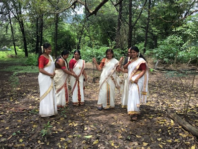 Five women dressed in traditional white saris with gold and red borders stand together in a wooded area, surrounded by green trees and plants. The ground is covered with fallen leaves and small branches. The women are smiling and engaged in conversation or interaction.