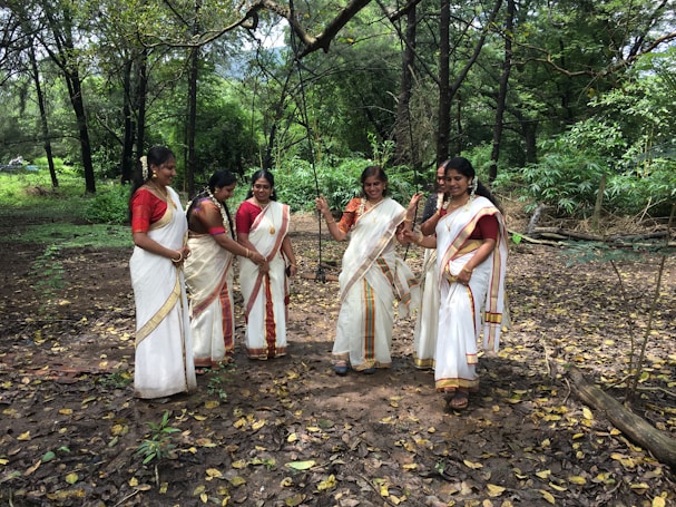 Five women dressed in traditional white saris with gold and red borders stand together in a wooded area, surrounded by green trees and plants. The ground is covered with fallen leaves and small branches. The women are smiling and engaged in conversation or interaction.