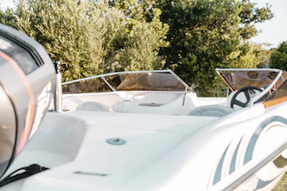 A close-up view of a sleek, modern boat with white and gray colors against a backdrop of lush green trees. The boat's interior features a steering wheel and tinted windshields.