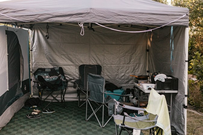Gray pop-up canopy at a campsite with gray sidewalls on two sides, camp chairs and a folding table with a stove on a green floor mat underneath