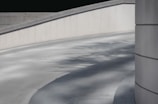 A concrete ramp with smooth curves, complemented by shadows of trees cast on its surface. The scene is framed by minimalist, modern architecture, featuring clean lines and a neutral color palette.