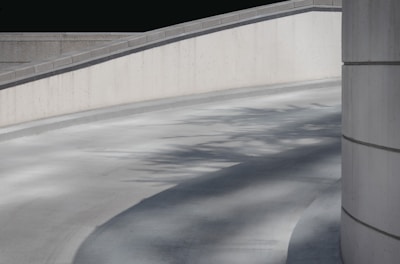 A concrete ramp with smooth curves, complemented by shadows of trees cast on its surface. The scene is framed by minimalist, modern architecture, featuring clean lines and a neutral color palette.