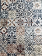 A collection of intricately designed ceramic tiles with various geometric and floral patterns. Each tile features unique motifs with combinations of blue, beige, and terracotta colors. The patterns range from symmetrical star shapes to more organic floral designs, providing a vintage and artistic appearance.