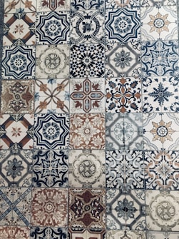 A collection of intricately designed ceramic tiles with various geometric and floral patterns. Each tile features unique motifs with combinations of blue, beige, and terracotta colors. The patterns range from symmetrical star shapes to more organic floral designs, providing a vintage and artistic appearance.