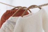 Three knit sweaters are hanging on wooden hangers. The sweaters are draped over a thick rope, showcasing their textures. The foreground sweater is cream-colored with a soft, fuzzy texture, while the middle sweater features cable knit details in a light beige hue. The rear sweater is a vibrant rust color.