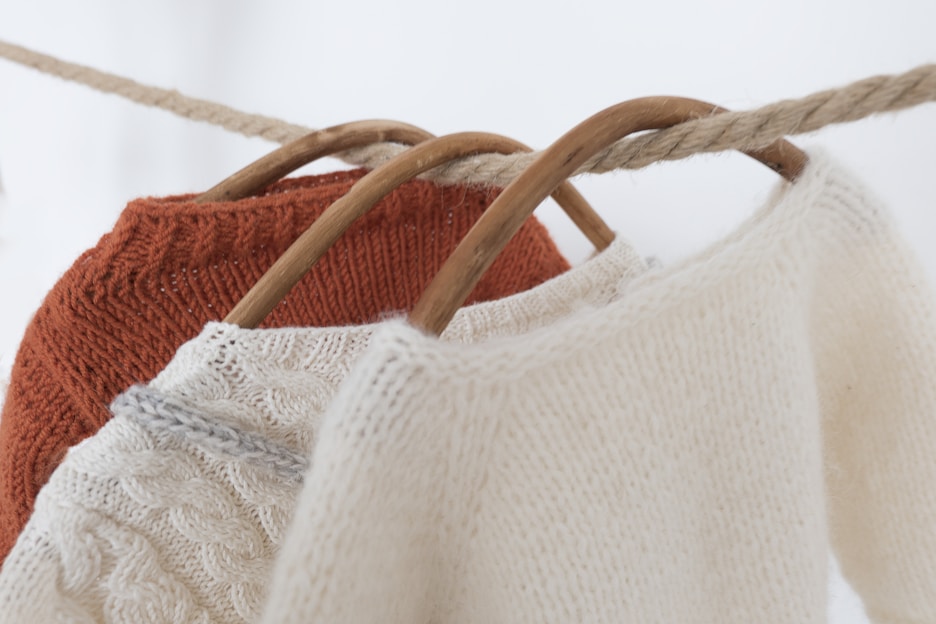 Three knit sweaters are hanging on wooden hangers. The sweaters are draped over a thick rope, showcasing their textures. The foreground sweater is cream-colored with a soft, fuzzy texture, while the middle sweater features cable knit details in a light beige hue. The rear sweater is a vibrant rust color.