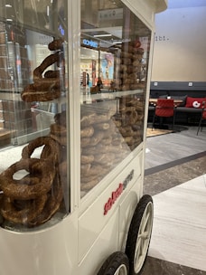 A glass display cart filled with stacks of simit, a type of bread, positioned in a mall setting. The cart has large wheels, and the surrounding area includes a seating section with red-cushioned chairs and a tiled floor.