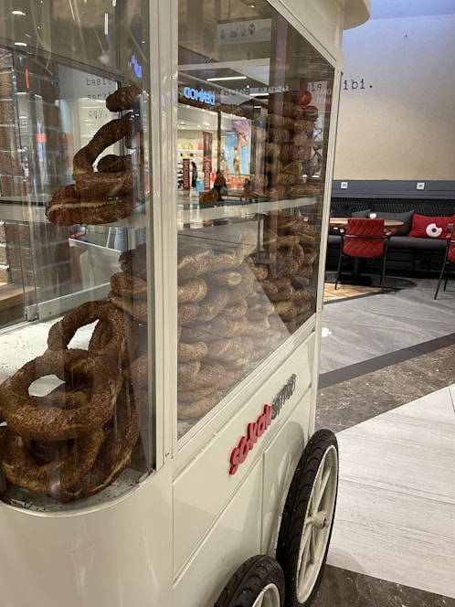 A glass display cart filled with stacks of simit, a type of bread, positioned in a mall setting. The cart has large wheels, and the surrounding area includes a seating section with red-cushioned chairs and a tiled floor.