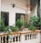 A small balcony features several potted green plants arranged on a light-colored balustrade, including leafy shrubs and small trees. A pair of glass-paned doors with metal grills and a colonial-style light fixture are part of the wall. The area conveys an old-world charm with the rustic style columns and slightly weathered walls.
