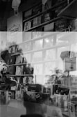 A black and white photo with a double exposure effect showing a person holding a record while standing in front of shelves filled with vinyl records displayed in a store. Various album covers are visible, including 'Lorde Pure' and 'Lana Del Rey.' Reflections and overlapping images create an artistic and abstract atmosphere.