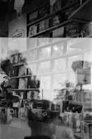 A black and white photo with a double exposure effect showing a person holding a record while standing in front of shelves filled with vinyl records displayed in a store. Various album covers are visible, including 'Lorde Pure' and 'Lana Del Rey.' Reflections and overlapping images create an artistic and abstract atmosphere.