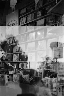 A black and white photo with a double exposure effect showing a person holding a record while standing in front of shelves filled with vinyl records displayed in a store. Various album covers are visible, including 'Lorde Pure' and 'Lana Del Rey.' Reflections and overlapping images create an artistic and abstract atmosphere.