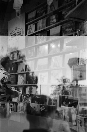 A black and white photo with a double exposure effect showing a person holding a record while standing in front of shelves filled with vinyl records displayed in a store. Various album covers are visible, including 'Lorde Pure' and 'Lana Del Rey.' Reflections and overlapping images create an artistic and abstract atmosphere.