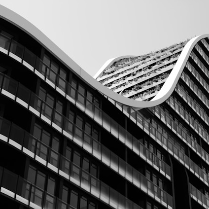 A modern architectural structure features a sleek, wavy design with a unique rooftop garden. The building's facade is composed of large glass windows with dark frames, creating a striking contrast against the light-colored wave. The vegetation on the rooftop adds a touch of nature to the otherwise minimalist and urban design.
