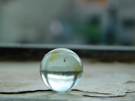A clear glass marble rests on a weathered surface, capturing and reflecting the soft ambient light. The background is softly blurred, suggesting an outdoor setting with muted natural tones.
