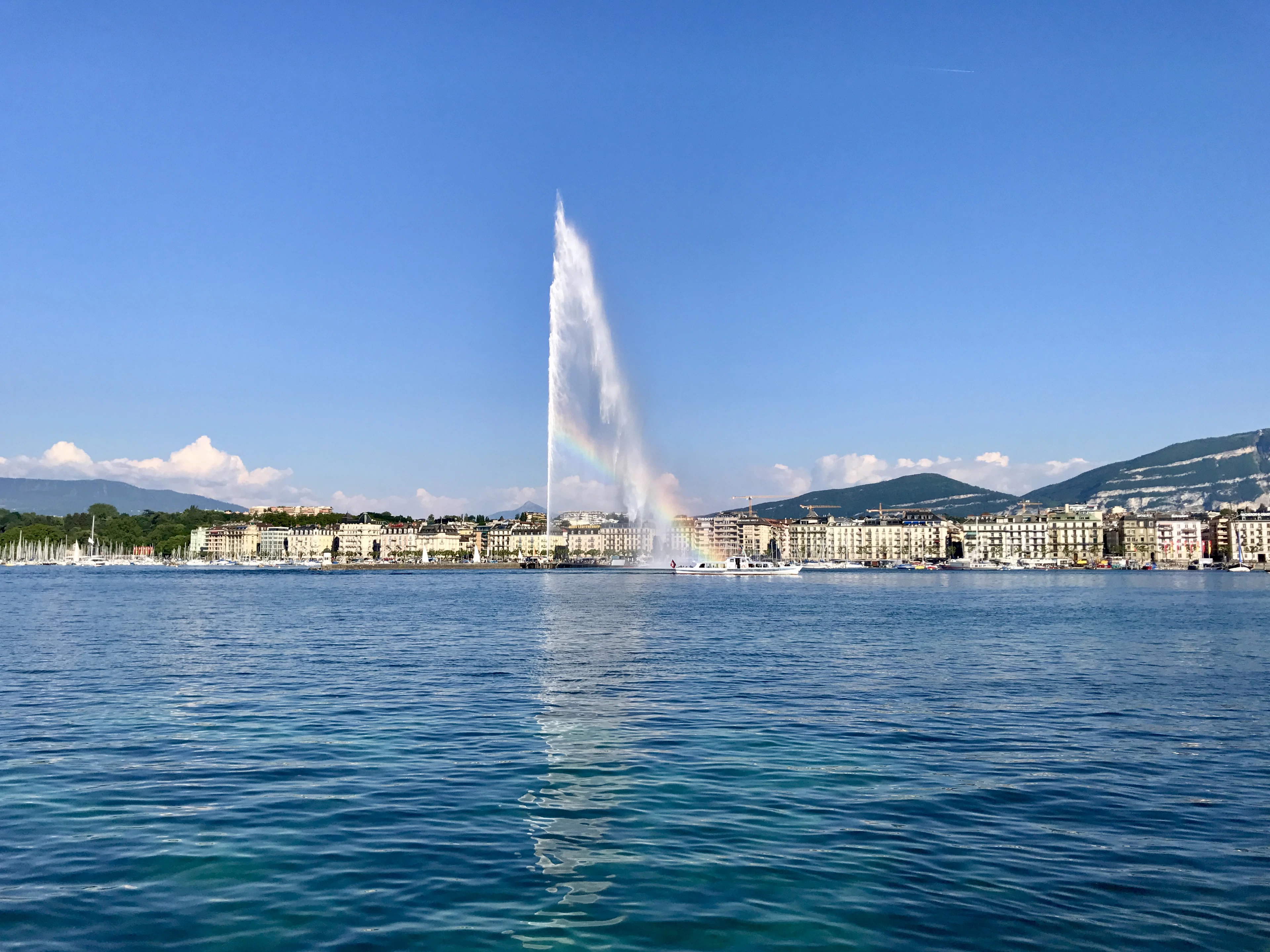 The Jet d'Eau fountain shooting a tall white plume of water from Lake Geneva, with the city's lakefront buildings and low green hills stretched across the horizon