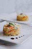 A carefully plated dish of mixed grains, including vibrant vegetables like orange carrots and potentially green peas, garnished with fresh green herbs. The dish is presented on a white plate with a striped pattern and accented by small drops of sauce.