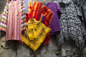 A collection of colorful, traditional garments hang on a stone wall. The assortment includes striped pants, a vivid red and orange poncho, a bright yellow and black poncho, a purple shirt, and a gray and black knitted cloak with geometric patterns. The stone wall provides a rustic backdrop to the vibrant textiles.
