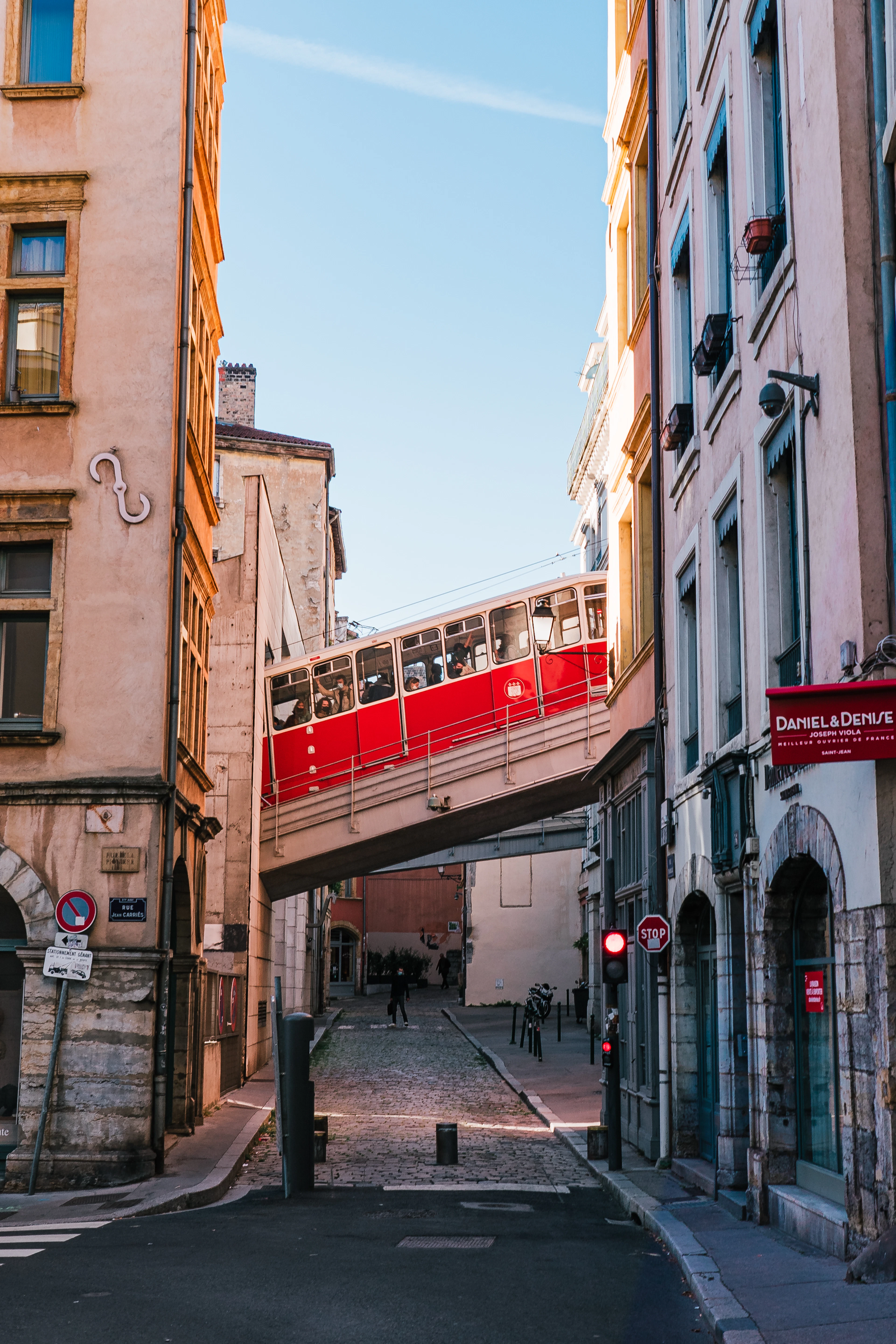 The red funicular carriage of Lyon's ficelle crossing a bridge high above a narrow street in Vieux Lyon, warm morning sun on the ochre facades on either side.