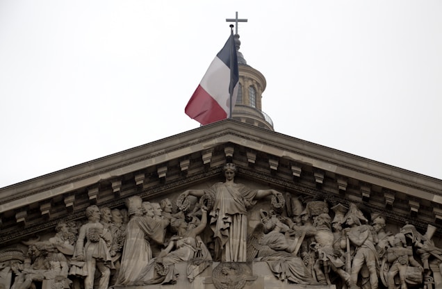 A neoclassical building with an elaborate sculptural frieze depicting historical figures and symbolic representations beneath a French flag. The sculptural work includes human figures dressed in classical and military attire, all positioned under a domed structure topped with a cross.