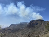 Thick smoke rises above a rugged mountain landscape with rocky cliffs. The sky is clear and blue, creating a stark contrast with the smoky haze.