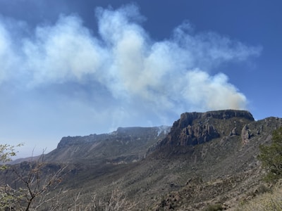 Thick smoke rises above a rugged mountain landscape with rocky cliffs. The sky is clear and blue, creating a stark contrast with the smoky haze.