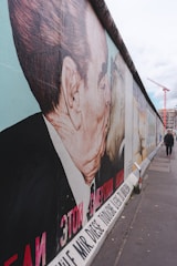A large mural on a concrete wall features two men kissing. The artwork is vibrant with significant text in multiple languages. The scene is outdoors, with a cloudy sky and a person walking in the distance. A construction crane and some urban structures can be seen far in the background.