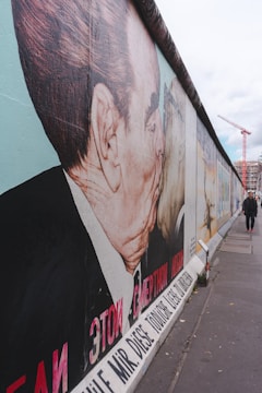 A large mural on a concrete wall features two men kissing. The artwork is vibrant with significant text in multiple languages. The scene is outdoors, with a cloudy sky and a person walking in the distance. A construction crane and some urban structures can be seen far in the background.