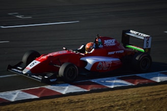 A red Formula racing car with the number 6 is moving swiftly around a track. The driver's helmet is visible, and the car is adorned with various sponsor logos including 'MRF TYRES' and 'ONET'. The track surface is dark with some red and white striped markings along the edges.