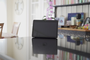 A digital tablet with a black screen is placed on a reflective surface in a well-lit room. In the background, there are shelves filled with books, a pink orchid, and various decorative items. The reflection of the tablet is visible on the shiny table surface.