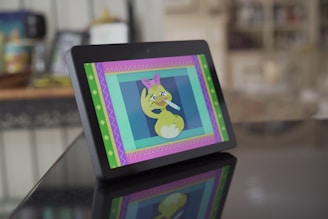 A tablet device is displayed on a reflective surface, showing a cartoon image of a sick, yellow duck with a pink bow, holding a thermometer in its mouth. The screen is framed by colorful, decorative borders. The background appears to be a domestic setting with household items slightly out of focus.