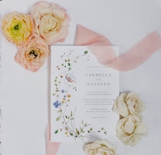 A wedding invitation card is surrounded by decorative flower arrangements. The card features floral designs and elegant typography. Beside it, there are clusters of pastel-colored flowers, including peach, pink, and cream roses. A soft pink ribbon lies across the surface, enhancing the romantic and delicate theme.