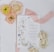 A wedding invitation card is surrounded by decorative flower arrangements. The card features floral designs and elegant typography. Beside it, there are clusters of pastel-colored flowers, including peach, pink, and cream roses. A soft pink ribbon lies across the surface, enhancing the romantic and delicate theme.