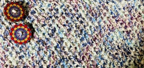 A close-up view of a knitted fabric featuring a textured pattern with a mix of colors including purple, blue, and beige. Two decorative circular buttons with intricate, colorful patterns are attached, providing a contrast against the muted tones of the fabric.