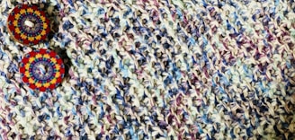 A close-up view of a knitted fabric featuring a textured pattern with a mix of colors including purple, blue, and beige. Two decorative circular buttons with intricate, colorful patterns are attached, providing a contrast against the muted tones of the fabric.
