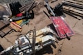 A collection of industrial tools and equipment scattered on a concrete surface. Several metal parts and pipes, including a red rectangular sign with white markings, rusty and worn, are visible. The area also contains a hydraulic component with hoses attached, along with various metal scraps and wooden planks.