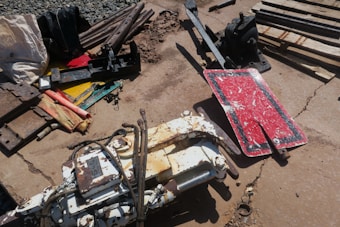 A collection of industrial tools and equipment scattered on a concrete surface. Several metal parts and pipes, including a red rectangular sign with white markings, rusty and worn, are visible. The area also contains a hydraulic component with hoses attached, along with various metal scraps and wooden planks.