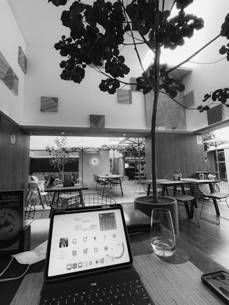 A black and white image depicts a modern indoor seating area with tables and chairs. A large tree is prominently positioned in the middle of the room, reaching up towards the ceiling. To the foreground, there's an open laptop displaying a home screen with app icons and a calendar. Next to it lies a glass with water, and the atmosphere is casual with elements of a caf&eacute; or lounge setting. There are decorative elements like wall panels and ambient lighting.