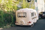 A small white caravan is parked in a residential area, surrounded by greenery and houses. The caravan features a vintage design and is close to a black vehicle. The scene is well-lit, suggesting it's daytime.