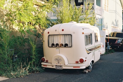 A small white caravan is parked in a residential area, surrounded by greenery and houses. The caravan features a vintage design and is close to a black vehicle. The scene is well-lit, suggesting it's daytime.