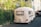 A small white caravan is parked in a residential area, surrounded by greenery and houses. The caravan features a vintage design and is close to a black vehicle. The scene is well-lit, suggesting it's daytime.