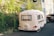 A small white caravan is parked in a residential area, surrounded by greenery and houses. The caravan features a vintage design and is close to a black vehicle. The scene is well-lit, suggesting it's daytime.