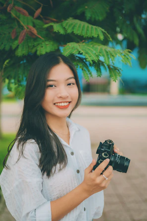 Portrait of a smiling young woman holding a camera in a sunlit urban setting.